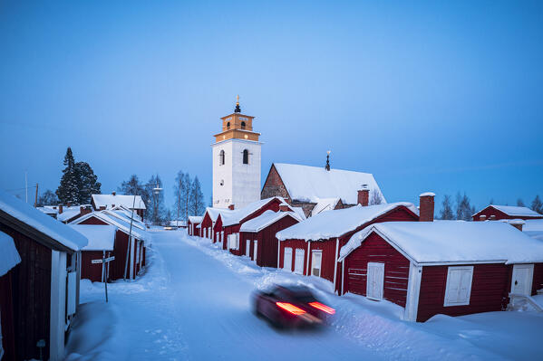 Car traveling on snowy road among traditional red cottages in the old town of Gammelstad at winter dusk, Lulea, Sweden