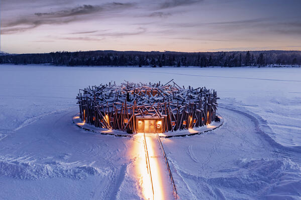 Illuminated circular building of the floating Arctic Bath Hotel and walkway covered with snow at dusk, Harads, Lapland, Sweden