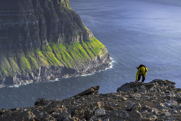 Man with backpack climbing the rocks of majestic cliff above the ocean, Vidoy Island, Faroe Islands (MR)