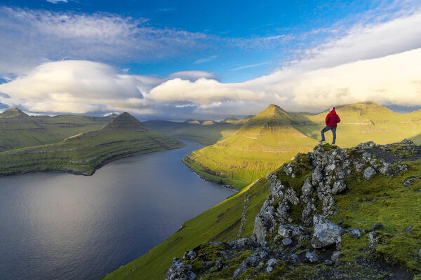 Hiker admiring the pristine water of a fjord standing on rocks at sunrise, Eysturoy Island, Faroe Islands (MR)