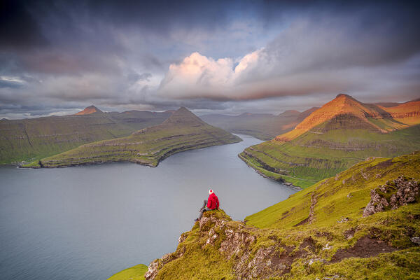 Hiker watching sunrise over the fjord sitting on mountain top, Eysturoy Island, Faroe Islands (MR)