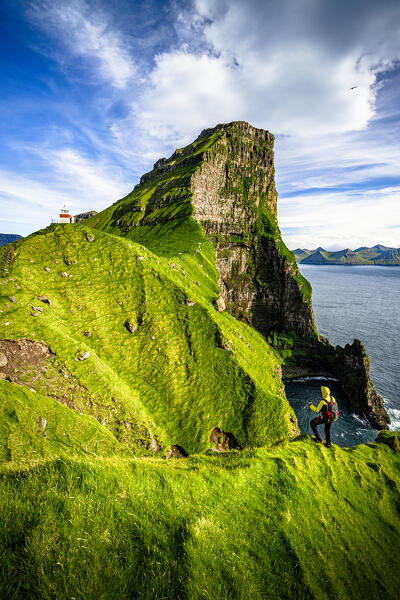 Hiker admiring Kallur lighthouse and Borgarin mountain peak standing on top of cliffs, Kalsoy island, Faroe Islands, Denmark