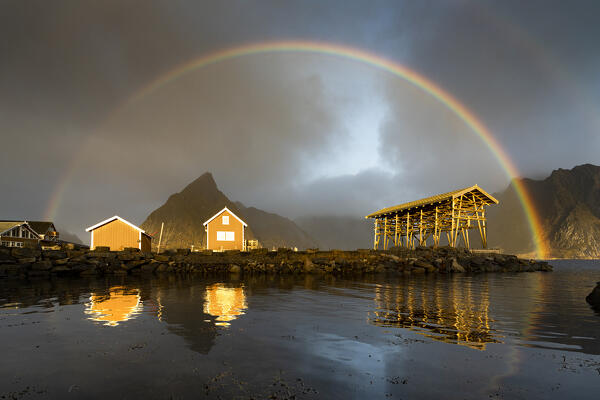 Fishermen cabins under a dramatic cloudy sky with  rainbow at dawn, Sakrisoy, Reine, Nordland county, Lofoten Islands, Norway
