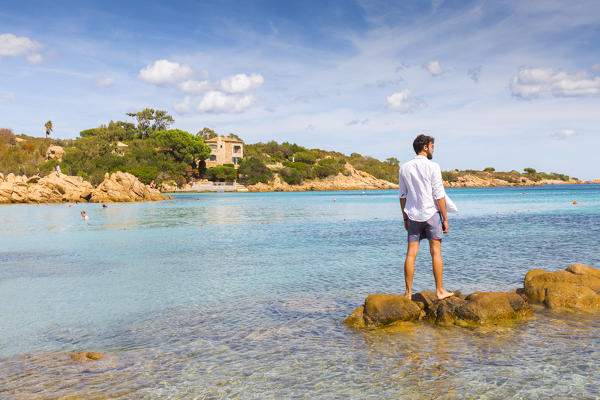 A young man admiring the view standing on rocks in Capriccioli beach, Arzachena Costa Smeralda, Olbia-Tempio province, Sardinia district, Italy