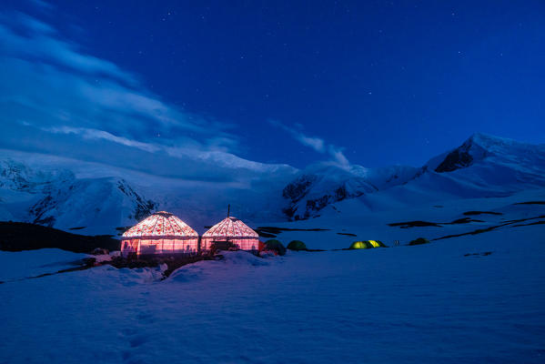 The yurts and tents of peak Lenin camp one illuminated at dusk with Peak Lenin massif in the background. Pamir, Kyrgyzstan, Central Asia. 