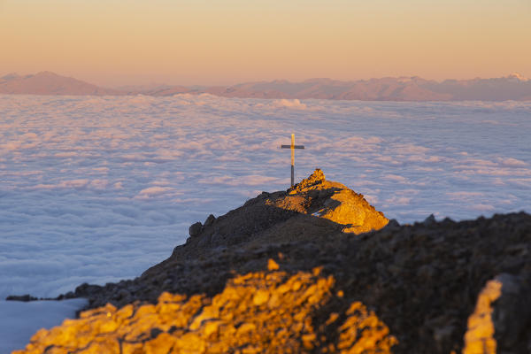 Summit cross of Latemar mount at sunrise over a cloudy carpet during summer. Schenon mount, Latemar group, Costalunga pass, Fassa valley, Pozza di Fassa, Trento district, Dolomites, Italy, Europe.