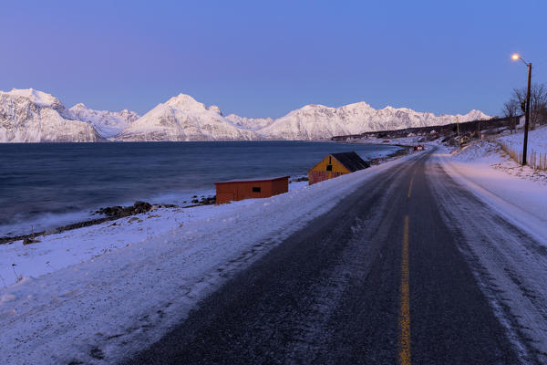 Road along the fjord illuminated by street lights during twilight. Hammarvika, Lyngenfjord, Lyngen Alps, Troms, Norway, Lapland, Europe.