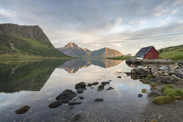 Typical rorbu and peaks reflected in the clear sea during midnight sun, Vengeren, Vagpollen, Lofoten Islands, Norway, Europe