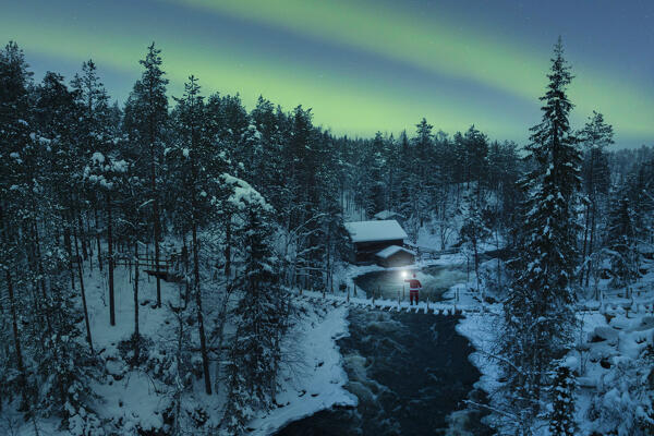 Santa Claus with lantern on the suspended bridge above the frozen rapids with the Northern Light in the sky, Myllykoski, Juuma, Oulanka National Park, Kuusamo, Lapland, Finland