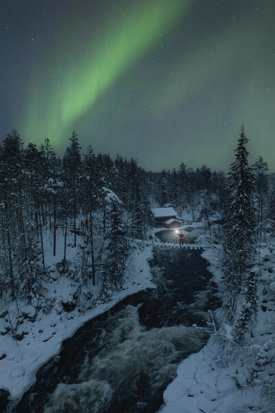 Santa Claus with lantern on the suspended bridge above the frozen rapids with the Northern Light in the sky, Myllykoski, Juuma, Oulanka National Park, Kuusamo, Lapland, Finland