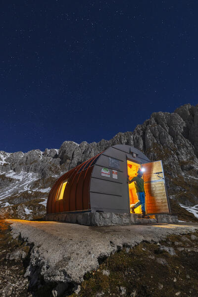 A trekker comes out of illuminated Città di Clusone Bivouac during night, Castione della Pesolana, Prealpi Orobie, Bergamo, Lombardy, Italy, Southern Europe