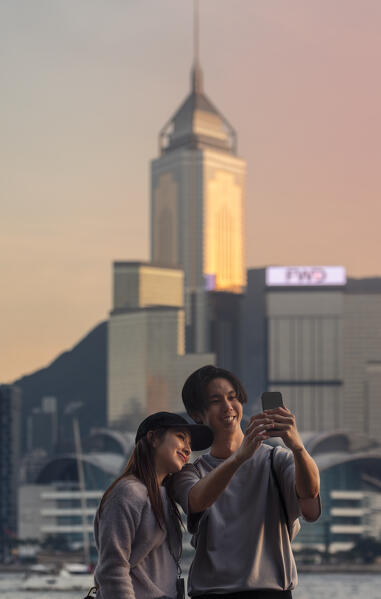 A couple takes a picture of themselves in front of cityscape during dusk viewed from Avenue of Stars, Tsim Sha Tsui, Kowloon, Hong Kong, China, Asia