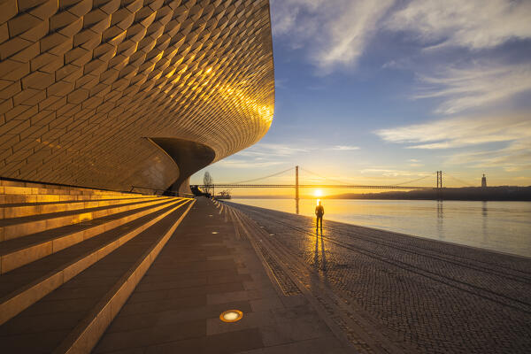 A tourist observes sunrise at Maat Museum with 25 April Bridge in the background, Belem, Lisbon, Portugal, Iberian peninsula, Western Europe