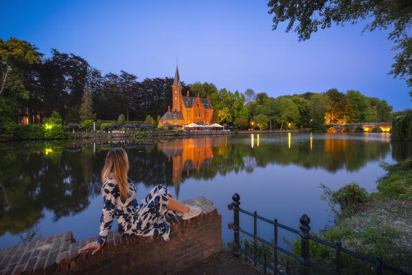 A tourist observes an illuminated Castillo de la Faille at Minnewater Park during dusk, Bruges, Belgium, Flanders, Western Europe
