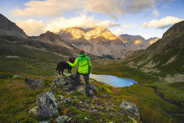 A hiker with his dog observe Lac Lavoir and mountains at sunset during summer from an elevated point of view, Provence Alps Cote d’Azur, Haute Alps, Vallee Etroite, France, Europe