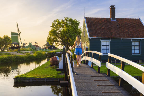 A tourist walks on a bridge in front of a typical house in Zaanse Schans during sunset in summer, Zaandam, Zaanstad, Netherlands, North Holland, Western Europe