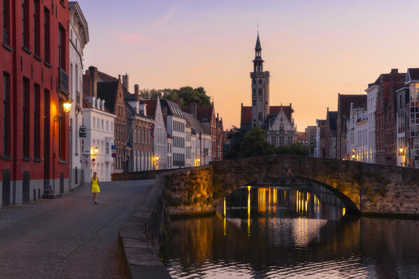 A tourist walks along the banks of the canal in front of Burghers Lodge at dusk, Bruges, Belgium, West Flanders, Flanders, Western Europe 