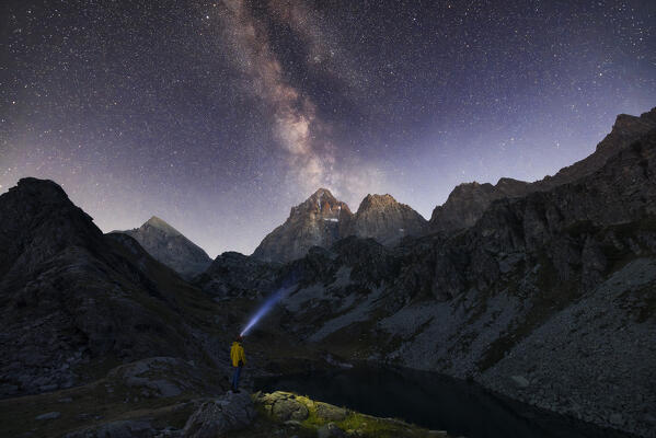 A trekker in front of Monviso over Fiorenza Lake observes Milky Way in the sky during night, Crissolo, Cuneo, Piedmont, Italy, Southern Europe