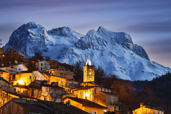 Winter dusk at the mountain village of Cerqueto with lights and illuminated bell tower and with Gran Sasso mountains in the background, Gran Sasso and Monti della Laga national park, Teramo province, Abruzzo, Italy