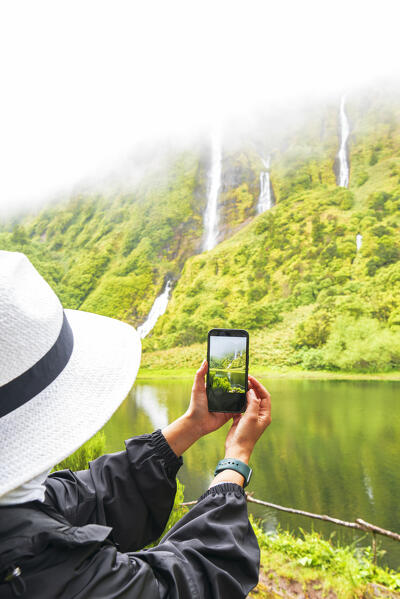 Rear view of a woman taking photo with the smarthphone of the Ribeira do Ferreiro waterfall with clouds, Poco Ribeira do Ferreiro (Alagoinha), Lajes das Flores municipality, Flores Island (Ilha das Flores), Azores archipelago, Portugal, Europe
