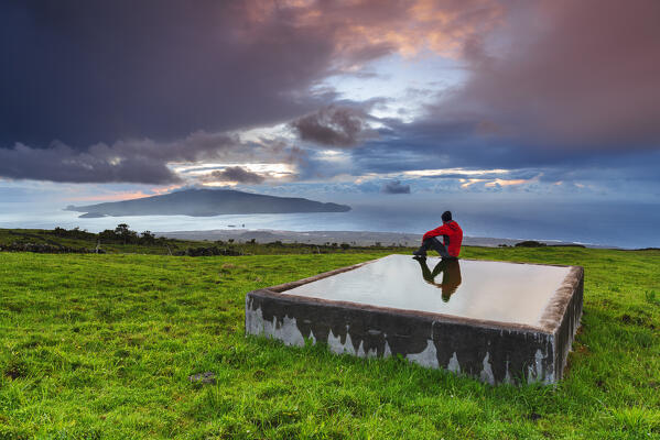 Man sits on the trough  and admires the view of Faial island from Pico island at sunset, Madalena municipality, Pico island (Ilha do Pico), Azores archipelago, Portugal, Europe