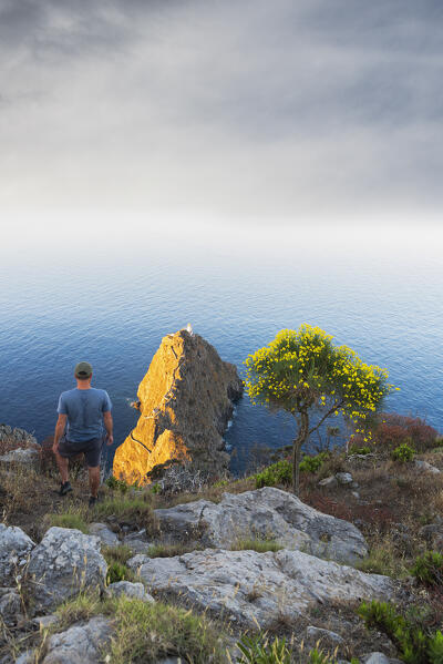 View from above of a man standing on top of the Monte della Guardia peak overlooking Punta della Guardia lighthouse at sunrise, Ponza island, Archipelago Pontino, Latina province, Latium, Italy