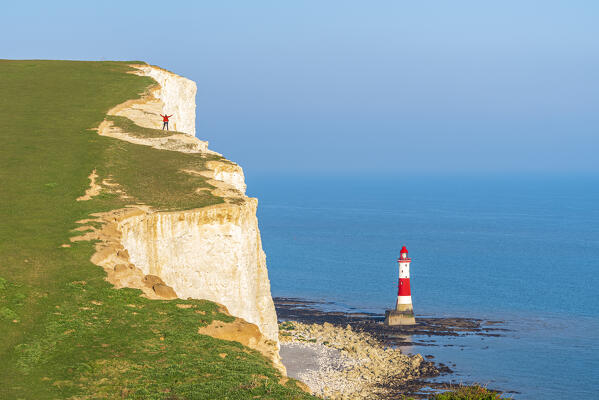 Hiker with arms raised stands on top of the white and chulky cliff overlooking Beachy Head lighthouse, Seven Sisters Country Park, South Downs National Park, Sussex, England, United Kingdom, Europe (MR)