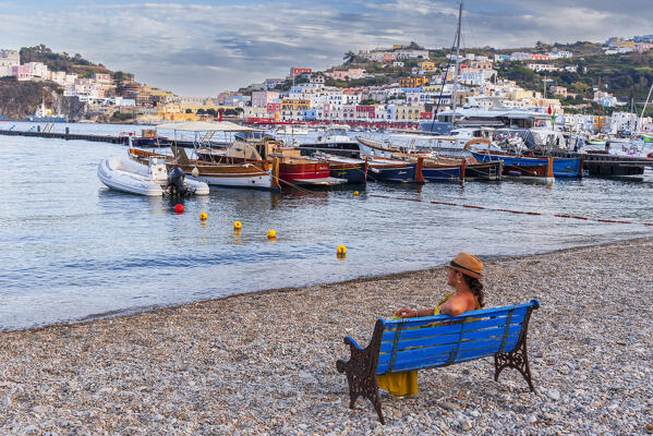 Beautiful woman with straw hat sit on a bench on the shingle beach of Ponza overlooking the marina and the colorful village at sunset, Ponza island, Pontine islands, Tyrrhenian Sea, Latina province, Latium, Italy, Europe
