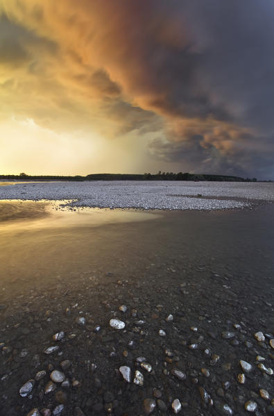 Po river park, Piedmont, Italy. Rocky beach on Po river banks, with stormy clouds coloured by the sunset lights.