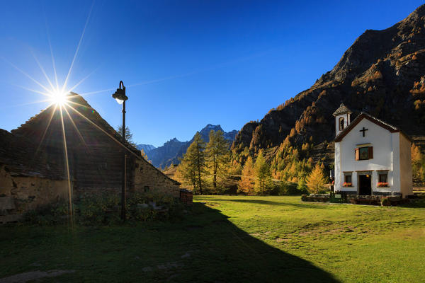 Sun flare behind one of the huts and the church of the alpine village of Crampiolo in autumn, Alpe Devero, Province of Verbano Cusio-Ossola, Piedmont, Italy, Europe