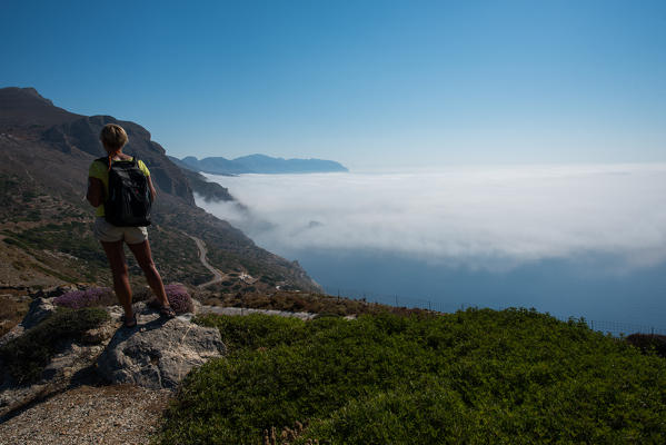 Amorgos island, Cyclades, Greece, Europe. A woman watching a seascape from the top of a hill. Near her flowers and in the sky the clouds.