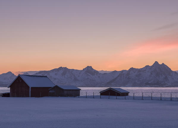 The orange sky at sunset on the wooden huts called Rorbu framed by frozen sea and snowy peaks in the municipality of Andoy Vesteralen district of Nordland county Norway Europe