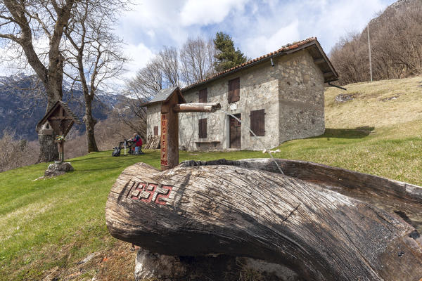 Couple of hikers sit on the benches outside the Nusieda Alta hut, in the foreground the large fountain carved into a wooden trunk. Monti del Sole, Dolomiti Bellunesi National Park, Belluno, Veneto, Italy