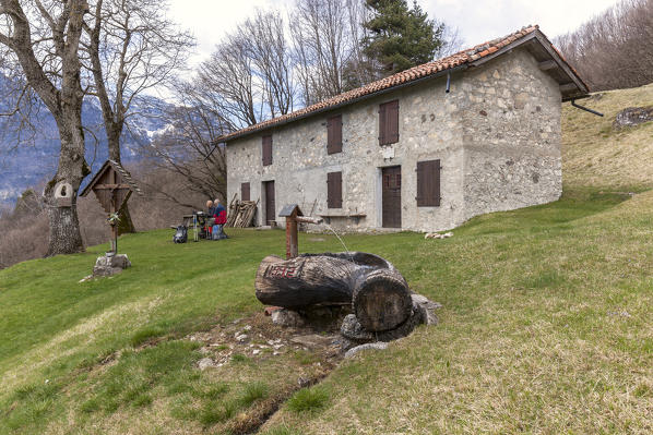 Couple of hikers sit on the benches outside the Nusieda Alta hut, in the foreground the large fountain carved into a wooden trunk. Monti del Sole, Dolomiti Bellunesi National Park, Belluno, Veneto, Italy