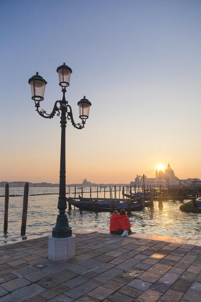two tourists admire the sunset behind the domes of the Basilica of Saint Mary of Health from the banks of the Grand Canal, Venice, Veneto, Italy